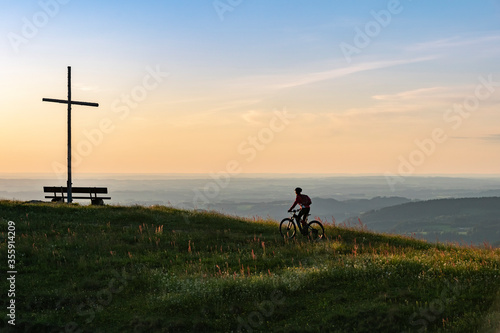 pretty senior woman riding her electric mountain bike in warm dawn sunlight below the summit cross of Salmas hight above Oberstaufen, Allgau Alps, Bavaria Germany 