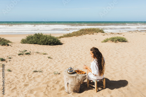 Young happy woman enjoying traditional moroccan mint tea outdoors on ocean sand beach in Morocco. View from behind.