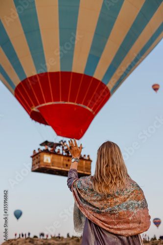 Beautiful blonde woman greets flying hot air balloon at dawn in Cappadocia