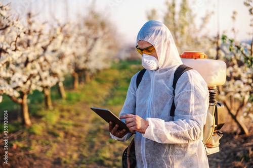 Fruit grower in protective uniform, mask and pollinator machine on his backs using tablet while standing in orchard.