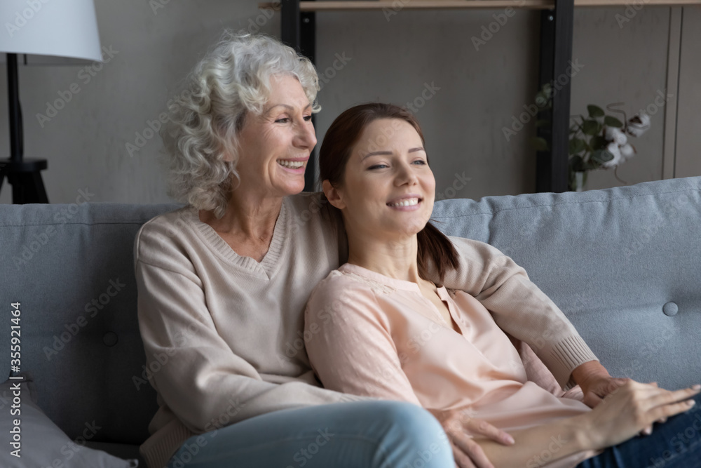 Smiling mature mother and adult daughter sit relax on couch at home hugging and cuddling, happy senior mom and grownup girl child rest on sofa embracing, look in distance dreaming together Smiling mature mother and adult daughter sit relax on couch at home hugging and cuddling, happy senior mom and grownup girl child rest on sofa embracing, look in distance dreaming together