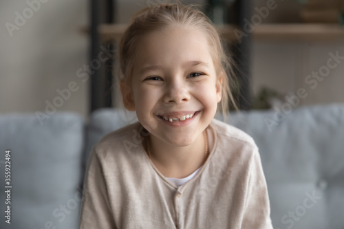 Close up headshot portrait of cute happy small preschooler girl with toothy smile look at camera posing, funny little child sit relax have fun on sofa in living room, feel optimistic and positive