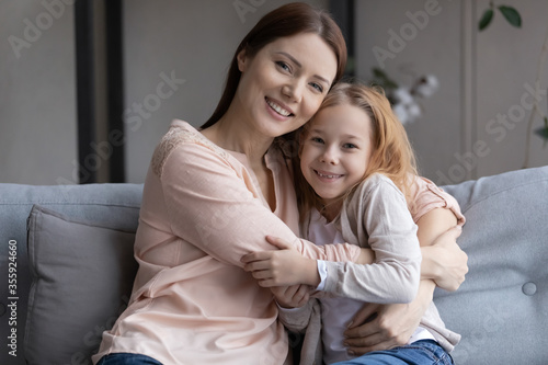 Portrait of happy young Caucasian mom and small preschooler daughter relax on sofa at home hugging cuddling, smiling mother and little girl child embrace, show love and care in family relationships