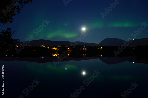 majestic aurora borealis, northern light over calm mirror lake at night with buildings in the background photo