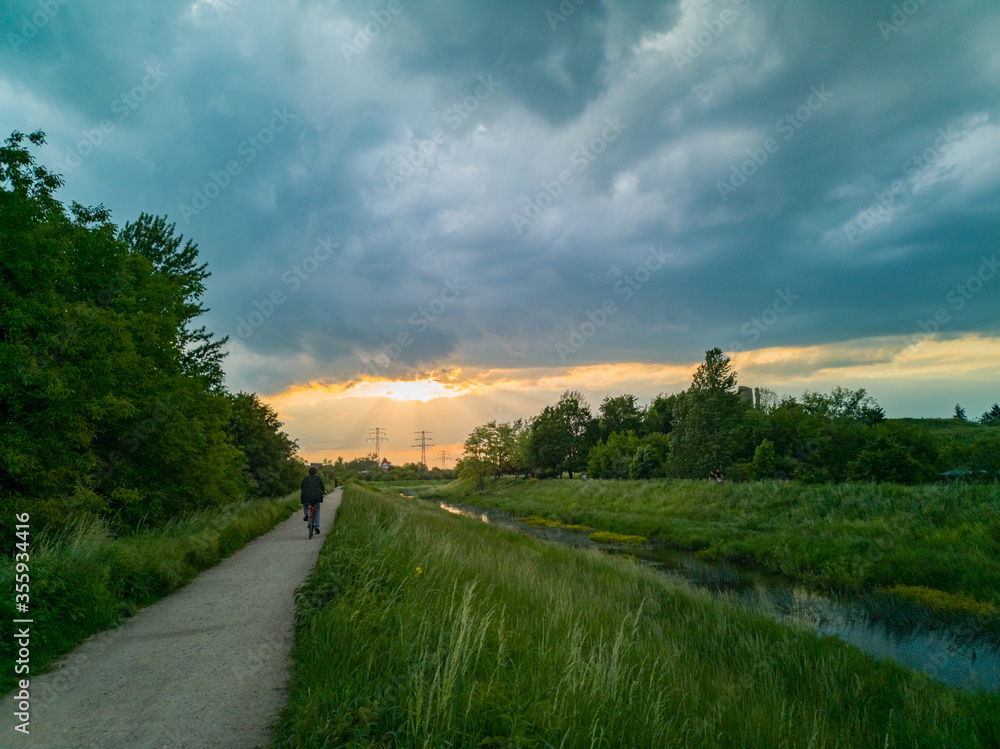 Fototapeta premium Beautiful cloudy sunset over long dark river in park with biker on path