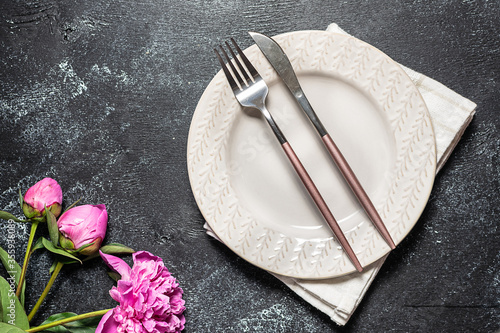 Plate,stylish cutlery set and pink peony flowers on black rustic table top view. Table setting.