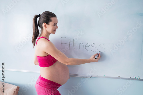Fototapeta Naklejka Na Ścianę i Meble -  A pregnant woman in sportswear writes the word fitness on a white marker board. A girl in the 3rd trimester goes in for sports with a bare tummy. Courses for expectant mothers.
