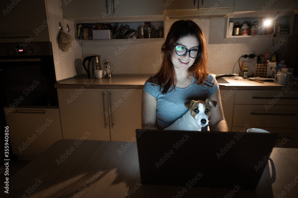 A smiling woman with glasses sits in the dark at a wireless computer in the kitchen with a puppy of Jack Russell Terrier on her knees. Girl with her little dog watching a movie on a laptop at home. A smiling woman with glasses sits in the dark at a wireless computer in the kitchen with a puppy of Jack Russell Terrier on her knees. Girl with her little dog watching a movie on a laptop at home.