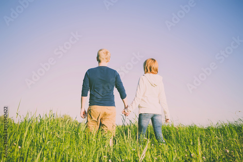 Young happy couple, man and woman, stand in grass with their backs to the camera. Great place for text on sky background. Sunny weather.