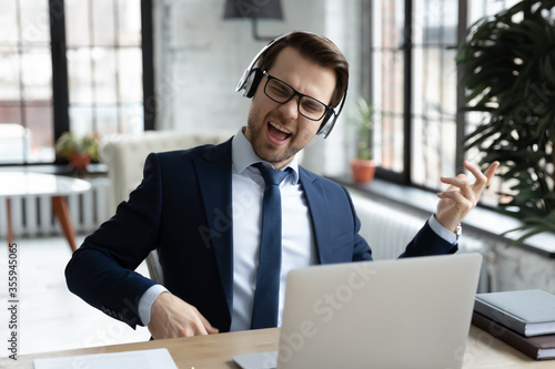 Overjoyed young Caucasian male office employee in suit wear headset listen to rock music at workplace, happy businessman in headphones relax have fun enjoy good quality audio, stress free concept