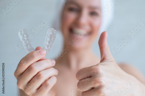 Orthodontic silicone transparent teeth aligner in female hands. A blurred woman with a towel on her head holds a removable retainer and shows a thumb up. Bracket for teeth whitening. Dental care.