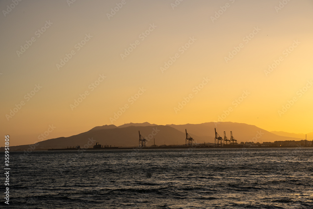 Summer sunset. Orange sky over the sea, mountains on the horizon and port container cranes.