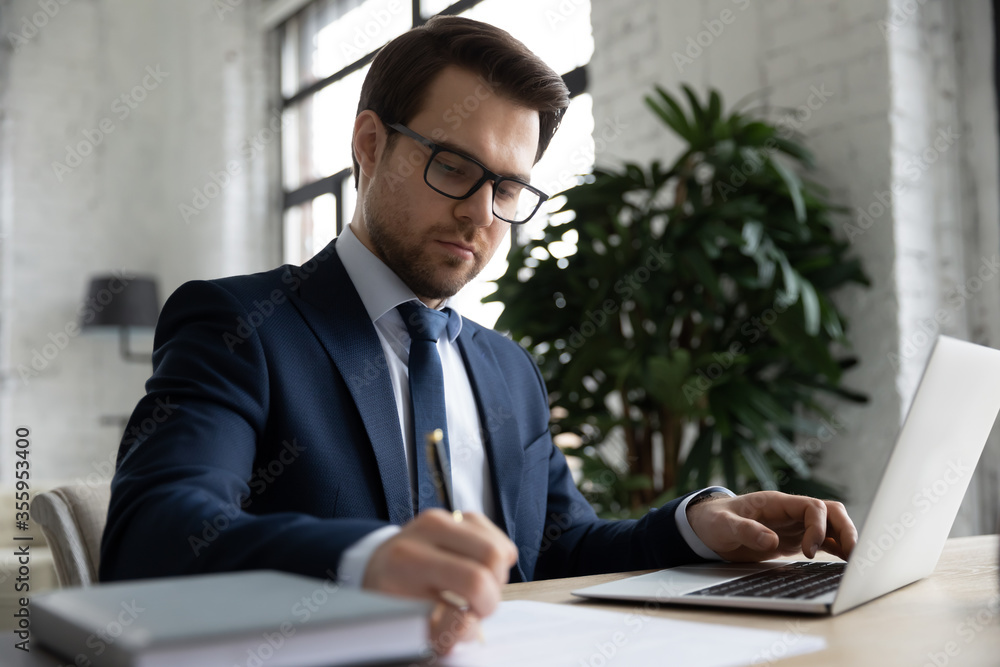 Concentrated Caucasian young businessman sit at desk in office work on laptop making notes, serious successful male employee in glasses busy writing preparing report at workplace, using computer Concentrated Caucasian young businessman sit at desk in office work on laptop making notes, serious successful male employee in glasses busy writing preparing report at workplace, using computer