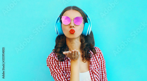 Portrait of attractive young woman in wireless headphones listening to music blowing red lips sending sweet air kiss wearing a pink sunglasses on colorful blue background