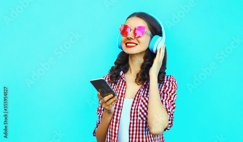 Portrait of young smiling woman with smartphone in wireless headphones listening to music wearing a pink sunglasses on colorful blue background