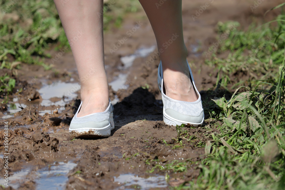 Female feet on a muddy path, shoes soiled in mud. Rural street with puddles after rain in summer, woman walking in the countryside