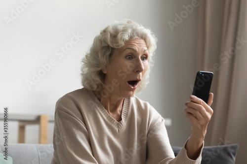 Shocked elderly 50s woman hold look at smartphone screen feel stunned by unexpected news online, amazed mature 60s female rest at home surprised by unbelievable message or text on cellphone device
