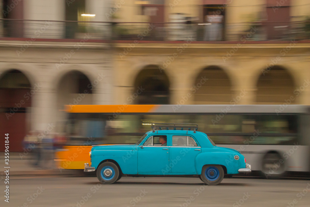Panning shot of classic blue car with blurred bus and building in background. Sunrise shot in Havana, Cuba
