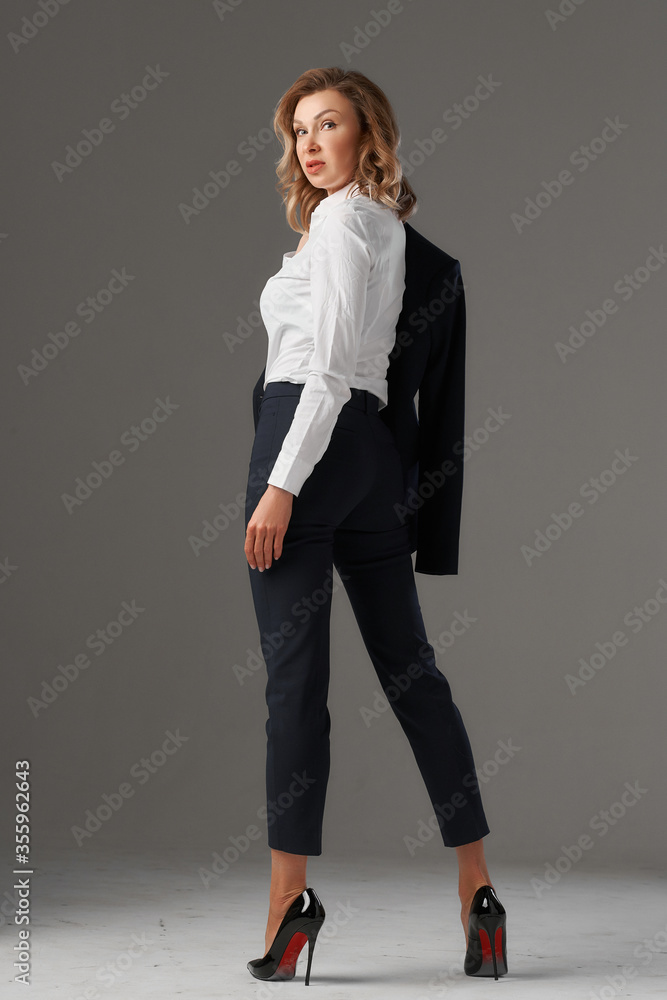 Stylish elegant middle aged woman with blonde hair wearing classical white blouse and black suit trousers and blazer standing and posing on gray background. Fashion studio portrait Stylish elegant middle aged woman with blonde hair wearing classical white blouse and black suit trousers and blazer standing and posing on gray background. Fashion studio portrait