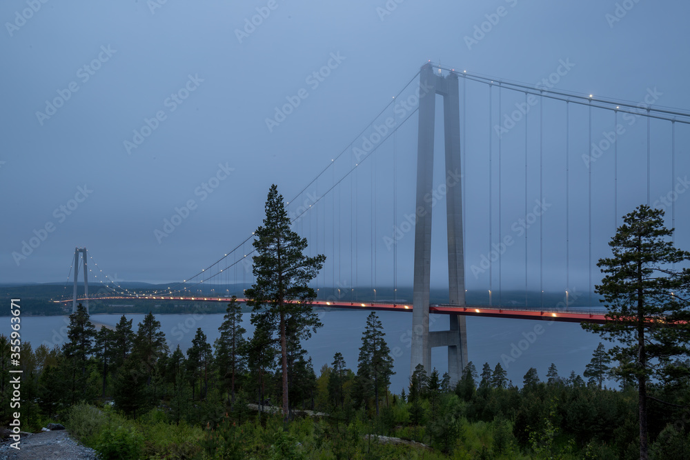 Picture of the illuminated Höga Kustenbron, a suspension bridge crossing the river Ångermanälven near Veda, Sweden taken on a late evening with low cloud cover around midsummer.
