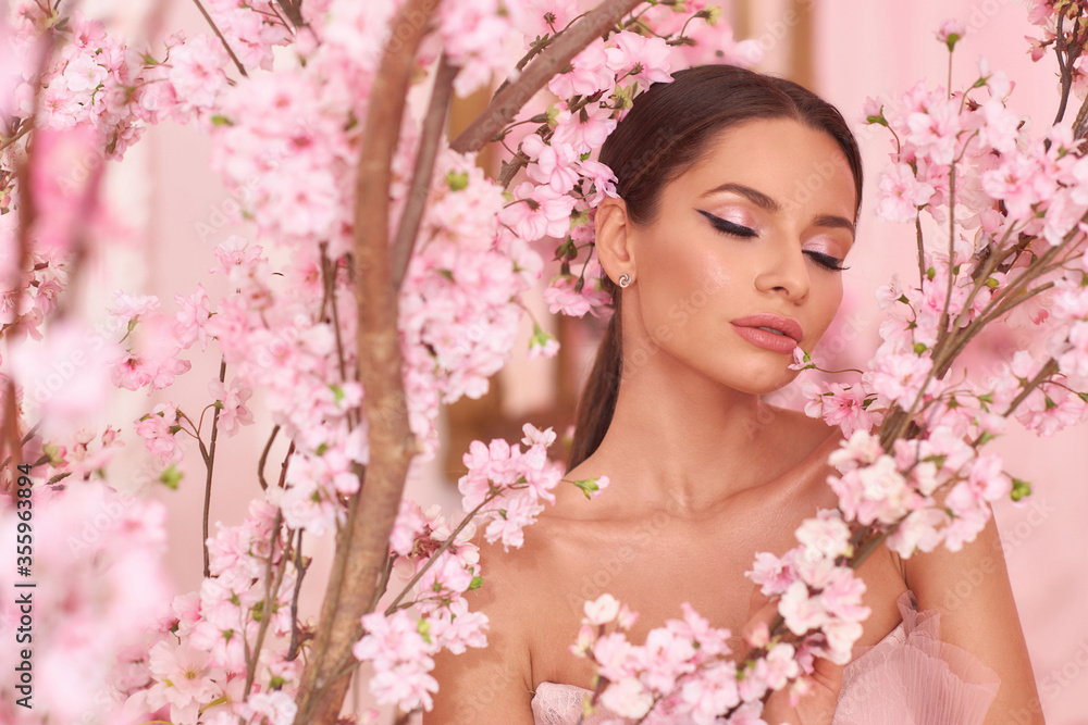 Closeup fashion spring face portrait of young beautiful caucasian woman with brunette hair in pony tail and perfect makeup looking through trees with pink flowers in blossom