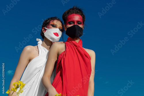 Two beautiful young brunette girls with creative bright makeup in tunics on a background of a field of yellow flowers and a blue sky. Two girls in medical masks. Healthcare