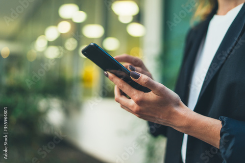Closeup photo business woman wearing suit, looking smartphone in hands. Open space loft office. Panoramic windows background. Wide mockup. Film effect