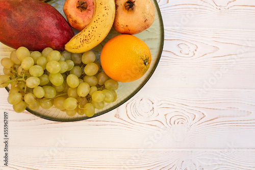 Juicy fruit grapes, banana, mango, pomegranate, oranges on a plate on a wooden white table. Copy space, flat lay