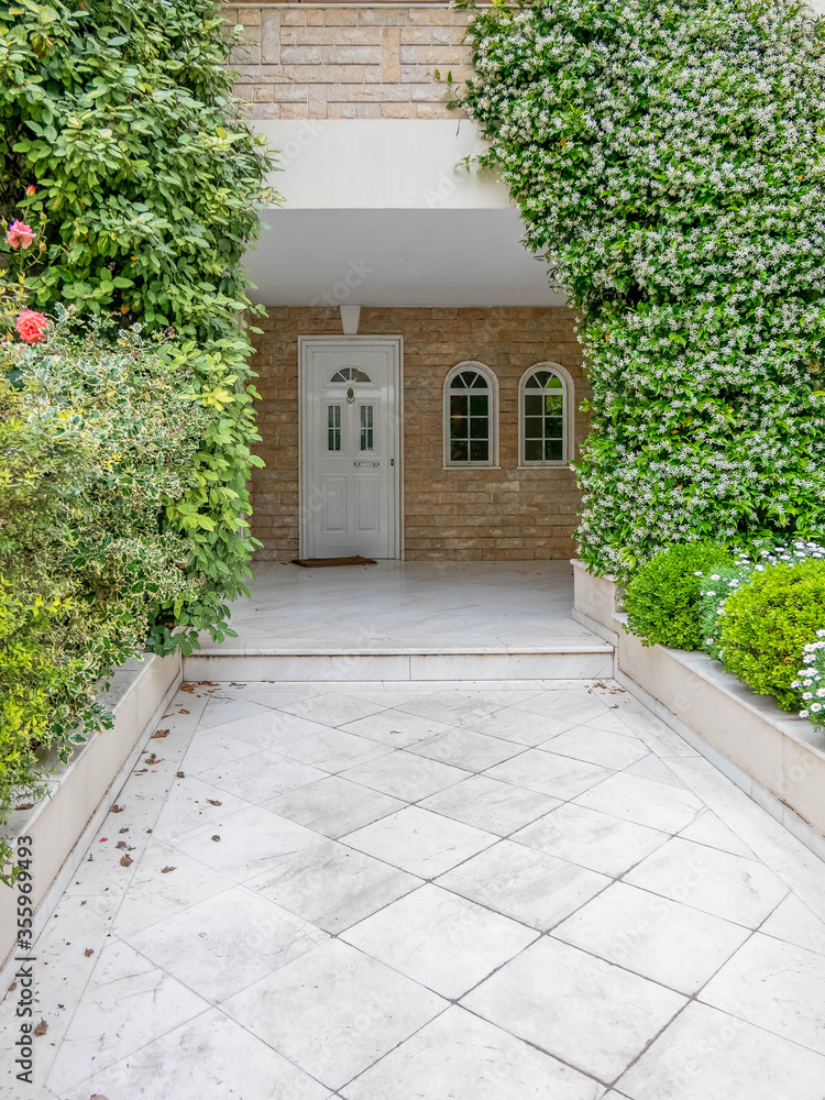 marble covered pathway to elegant house entrance white door  with plenty of foliage and flowers