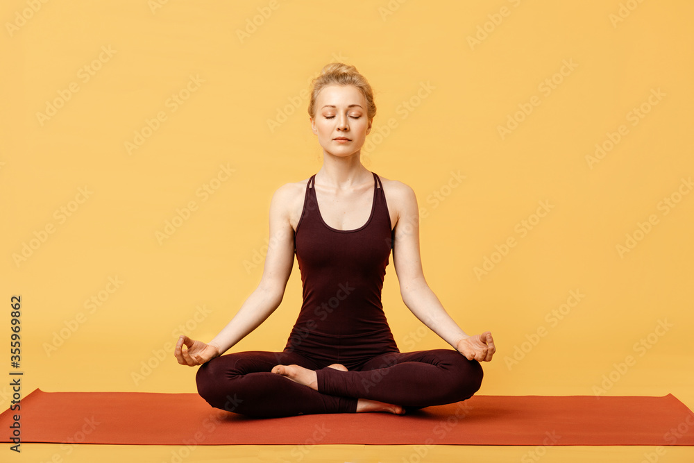 Young blond woman meditating, doing yoga lotus padmasana pose and asana. Fitness girl enjoying yoga indoors in sport clothes on yellow background, isolated. Stress-free yogic meditation practice Young blond woman meditating, doing yoga lotus padmasana pose and asana. Fitness girl enjoying yoga indoors in sport clothes on yellow background, isolated. Stress-free yogic meditation practice