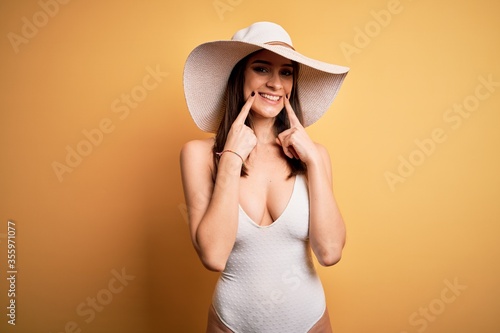 Young beautiful brunette woman on vacation wearing swimsuit and summer hat Smiling with open mouth, fingers pointing and forcing cheerful smile