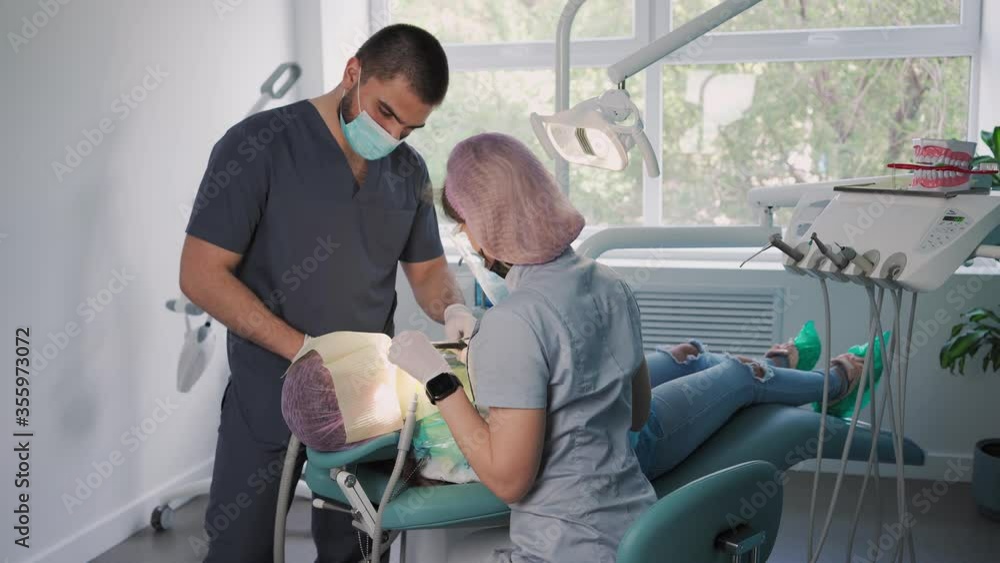 Beautiful Woman Dentist in a Protective Shield and Face Mask with the Help of an Male Assistant and Medical Equipment Works with a Female Patient in Dental Office. Healthcare and Medicine Concept
