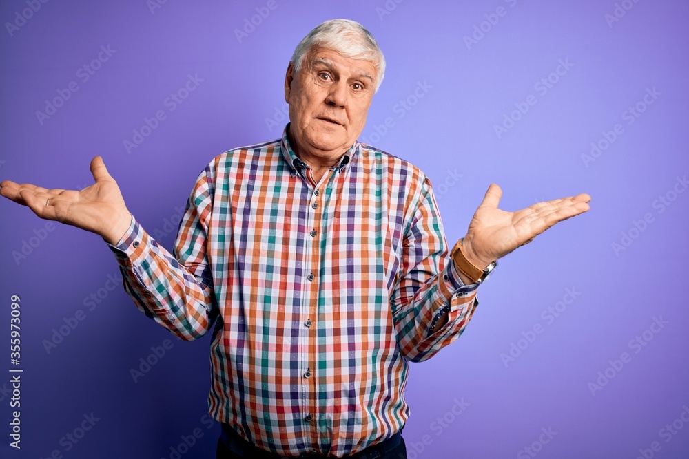 Naklejka premium Senior handsome hoary man wearing casual colorful shirt over isolated purple background clueless and confused expression with arms and hands raised. Doubt concept.