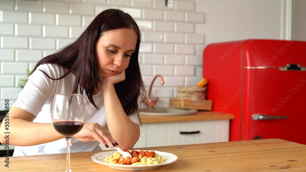 Young woman picks with fork in dish. Adult female sits thoughtfully in kitchen over plate of food. Young woman picks with fork in dish. Adult female sits thoughtfully in kitchen over plate of food.