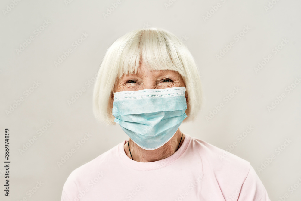 Portrait of happy female senior patient wearing medical protective mask looking at camera and smiling while standing against grey background Portrait of happy female senior patient wearing medical protective mask looking at camera and smiling while standing against grey background
