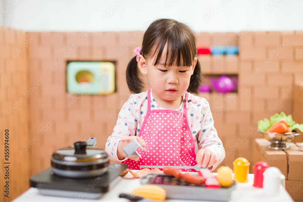 Fototapeta premium toddler girl pretend play food preparing role against cardboard blocks kitchen background