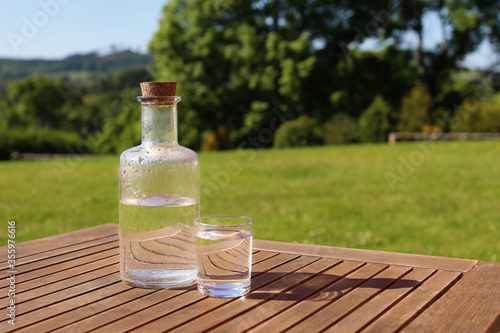 Bottle and glass of water on wooden table with chair. Blurred green trees and hill tops background. Outdoor breakfast in countryside. Picnic, garden party snack, relaxation concept. No people.