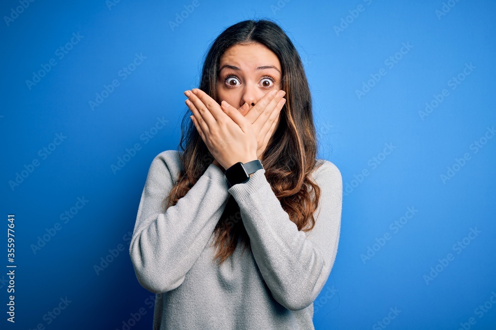 Young beautiful brunette woman wearing casual sweater standing over blue background shocked covering mouth with hands for mistake. Secret concept. Young beautiful brunette woman wearing casual sweater standing over blue background shocked covering mouth with hands for mistake. Secret concept.