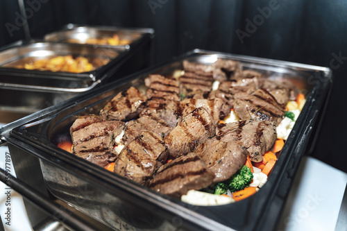Stainless hotel pans on food warmers with various meals. Grilled beef steaks laying on vegetable. Self-service buffet table. Celebration,party,wedding