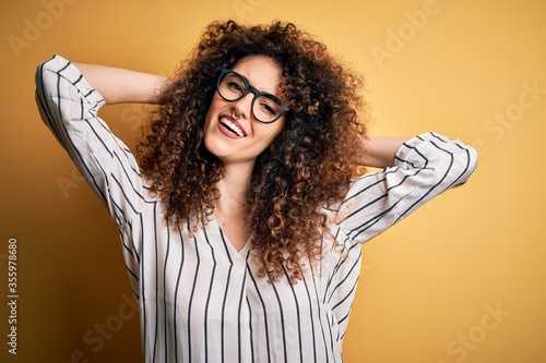 Young beautiful woman with curly hair and piercing wearing striped shirt and glasses relaxing and stretching, arms and hands behind head and neck smiling happy