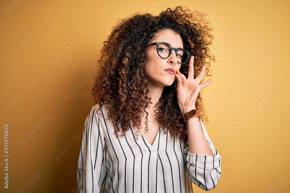Young beautiful woman with curly hair and piercing wearing striped shirt and glasses mouth and lips shut as zip with fingers. Secret and silent, taboo talking Young beautiful woman with curly hair and piercing wearing striped shirt and glasses mouth and lips shut as zip with fingers. Secret and silent, taboo talking