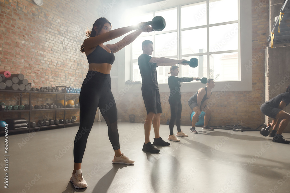 Total commitment. Full length shot of young cheerful athletic woman lifting a weight while having workout at industrial gym. Group training, teamwork concept Total commitment. Full length shot of young cheerful athletic woman lifting a weight while having workout at industrial gym. Group training, teamwork concept