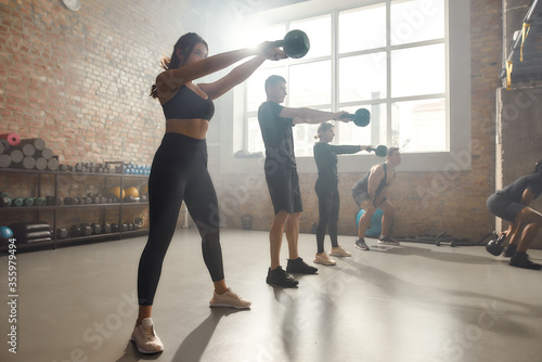 Total commitment. Full length shot of young cheerful athletic woman lifting a weight while having workout at industrial gym. Group training, teamwork concept