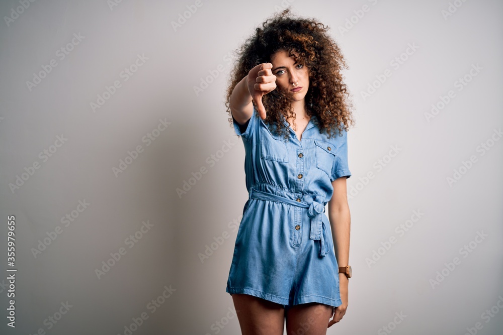 Young beautiful woman with curly hair and piercing wearing casual denim dress looking unhappy and angry showing rejection and negative with thumbs down gesture. Bad expression. Young beautiful woman with curly hair and piercing wearing casual denim dress looking unhappy and angry showing rejection and negative with thumbs down gesture. Bad expression.