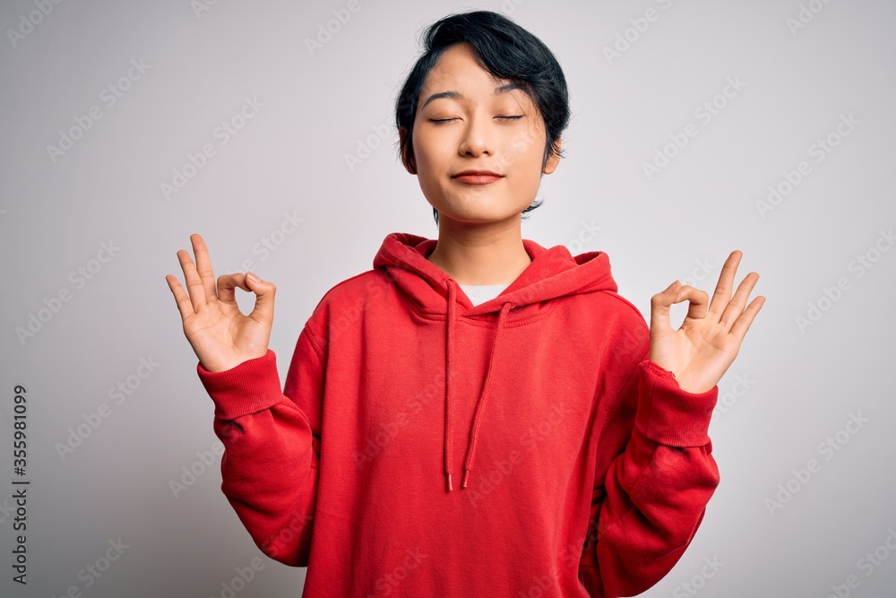 Young beautiful asian girl wearing casual sweatshirt with hoodie over white background relax and smiling with eyes closed doing meditation gesture with fingers. Yoga concept. Young beautiful asian girl wearing casual sweatshirt with hoodie over white background relax and smiling with eyes closed doing meditation gesture with fingers. Yoga concept.