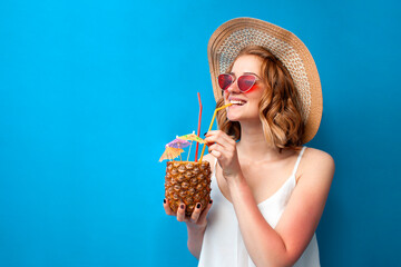 girl in a sun hat and glasses drinks a tropical pineapple cocktail on a blue isolated background, a woman with an exotic drink is relaxing in the resort, summer concept