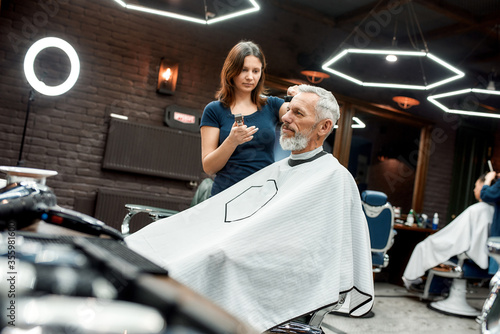 Making stylish hairstyle in barber shop. Side view of young professional barber girl using hair clipper. She is making new haircut for elegant mature man sitting in armchair in the front of mirror