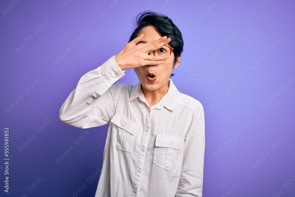 Young beautiful asian girl wearing casual shirt and glasses standing over purple background peeking in shock covering face and eyes with hand, looking through fingers with embarrassed expression. Young beautiful asian girl wearing casual shirt and glasses standing over purple background peeking in shock covering face and eyes with hand, looking through fingers with embarrassed expression.