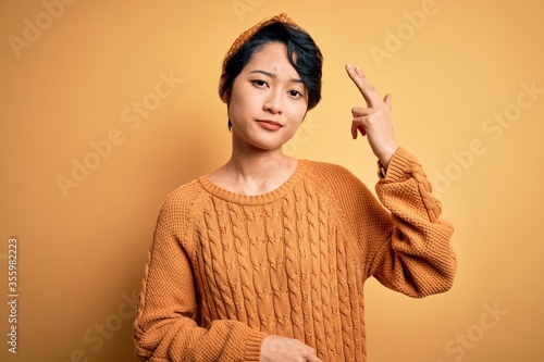 Young beautiful asian girl wearing casual sweater and diadem standing over yellow background Shooting and killing oneself pointing hand and fingers to head like gun, suicide gesture.