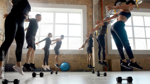 Let the bodies hit the floor. Group of sportive people jumping, warming up before having workout with dumbbells at industrial gym photo